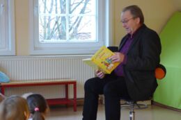 Bürgermeister Veit Künzelmann sitzt auf einem Stuhl in einem Grupenraum und liest Kindergartenkindern, die auf dem Boden vor ihm sitzen, eine Geschichte vor.